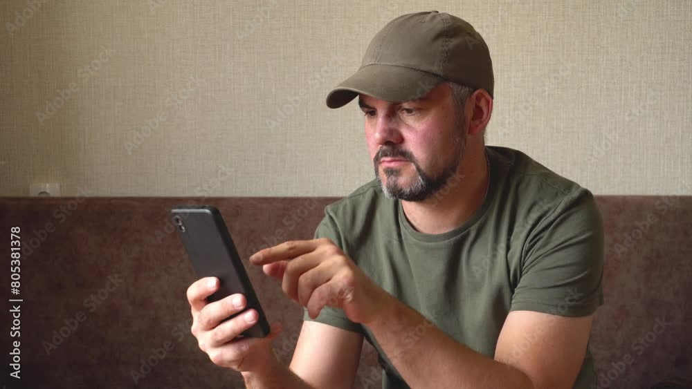 A man with a beard. Holding a black smartphone in his hand. Presses his finger on the screen. Thoughtful look. Green baseball cap and T-shirt. Military. In room. Sitting on a brown sofa against the ba A man with a beard. Holding a black smartphone in his hand. Presses his finger on the screen. Thoughtful look. Green baseball cap and T-shirt. Military. In room. Sitting on a brown sofa against the ba
