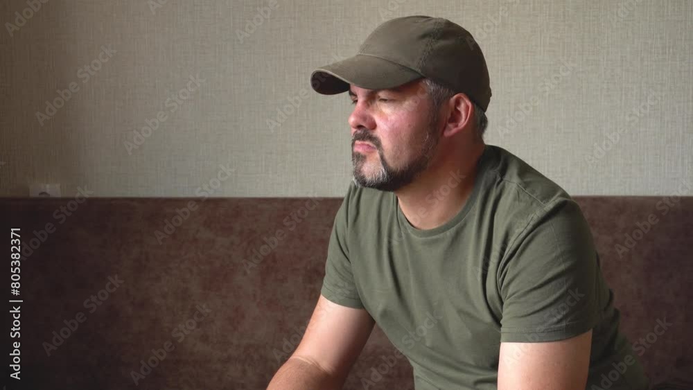 The man looks thoughtfully into the distance. Bearded face. Green baseball cap and clothes. Grey hair. In room. Reflections. Military. Close-up. The man looks thoughtfully into the distance. Bearded face. Green baseball cap and clothes. Grey hair. In room. Reflections. Military. Close-up.