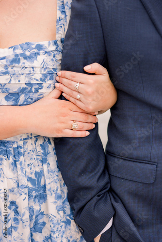 A close-up of a woman's hands that are wrapped around a man. He wears a navy blue suit jacket, and she is in a blue floral dress. 