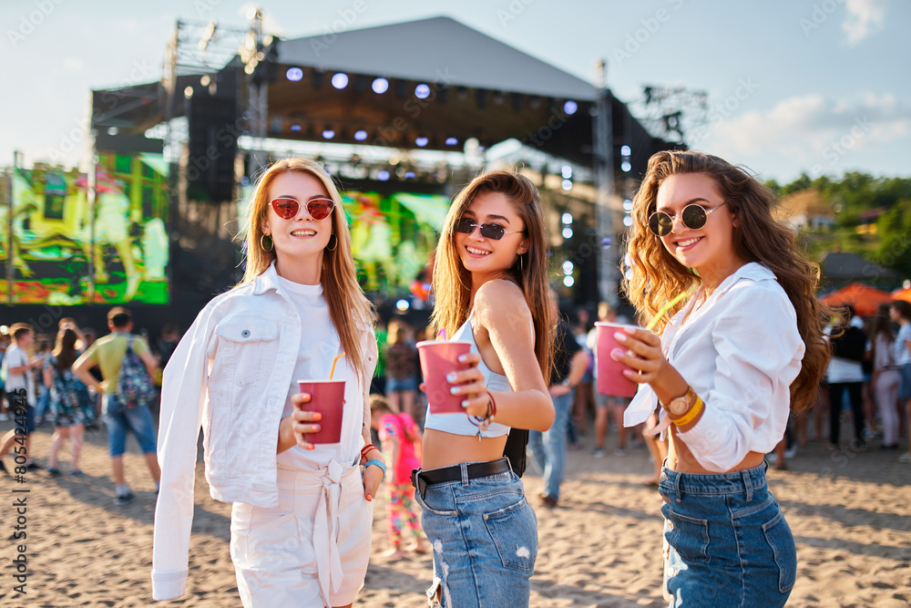 Group of girls at summer beach music festival, raising glasses in toast, enjoying sunny day with live concert in background. Smiling females celebrate, dance with drinks in hand. Group of girls at summer beach music festival, raising glasses in toast, enjoying sunny day with live concert in background. Smiling females celebrate, dance with drinks in hand.