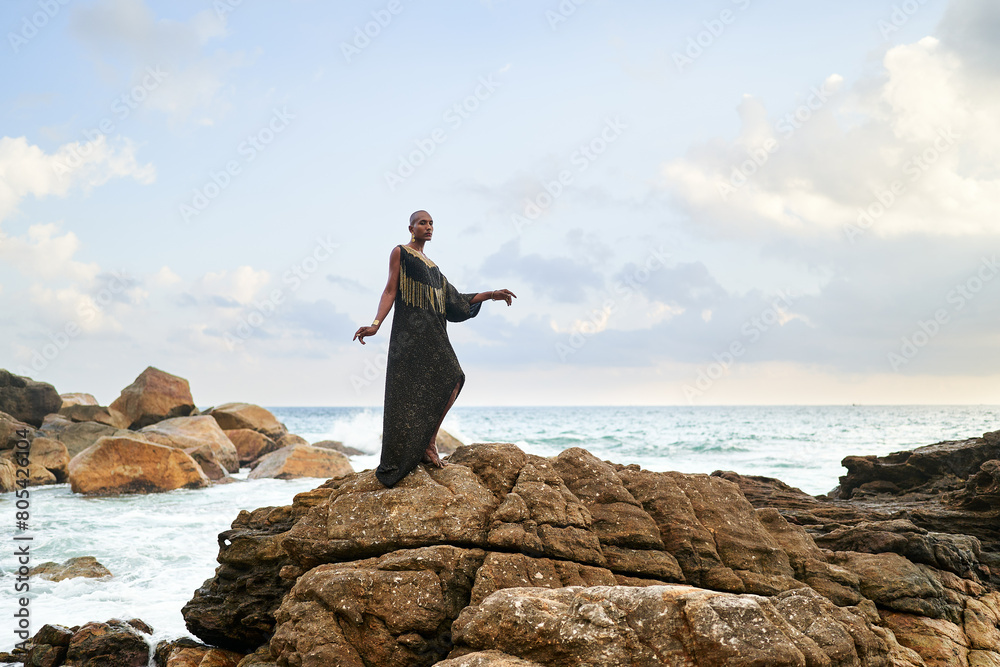 Non-binary black godlike person poses gracefully standing on rocks in ocean. Trans ethnic fashion model in posh dress and jewellery on rocky beach by storm . Lgbtq. Pride month Non-binary black godlike person poses gracefully standing on rocks in ocean. Trans ethnic fashion model in posh dress and jewellery on rocky beach by storm . Lgbtq. Pride month