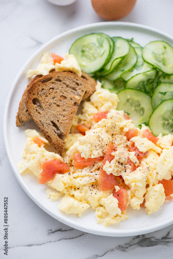 Scrambled eggs with smoked salmon, toasts and cucumber salad, vertical shot on a white marble background, middle close-up Scrambled eggs with smoked salmon, toasts and cucumber salad, vertical shot on a white marble background, middle close-up
