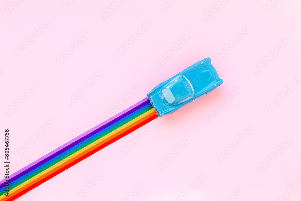 Blue toy car, rainbow ribbon flag trail road, pink background. June gay parade, proud month, coming out day, human rights and tolerance concept. Minimal pop art composition. Flat lay, top view Blue toy car, rainbow ribbon flag trail road, pink background. June gay parade, proud month, coming out day, human rights and tolerance concept. Minimal pop art composition. Flat lay, top view