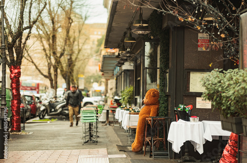A cozy teddy bear sits on a wooden table outside a charming restaurant, surrounded by the bustle of a lively street