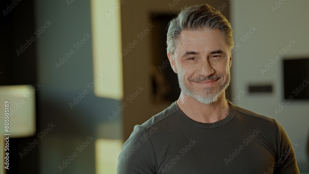 Fototapeta premium Portrait of a cheerful mature man with a gray beard smiling indoors. Confident fit muscular mid adult older male in 50s.