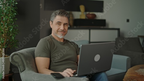 Confident mature man using laptop and smartphone, sitting in modern living room. Businessman working at home with computer, smiling. Happy mid adult man. Entrepreneur managing business online. © nyul
