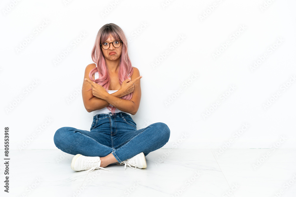 Young mixed race woman with pink hair sitting on the floor isolated on white background pointing to the laterals having doubts Young mixed race woman with pink hair sitting on the floor isolated on white background pointing to the laterals having doubts