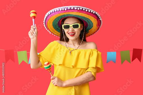 Happy young woman in Mexican sombrero hat and with maracas on red background