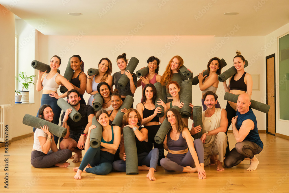 A large group of people are posing for a photo with yoga mats. Scene is positive and energetic, as everyone is smiling and holding their mats. The idea of the image is to capture a moment of camarader