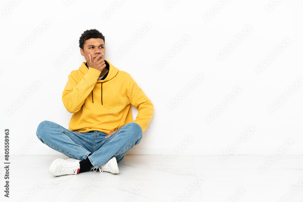 Young African American man sitting on the floor isolated on white background having doubts and with confuse face expression Young African American man sitting on the floor isolated on white background having doubts and with confuse face expression