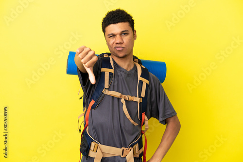 Young mountaineer African American man with a big backpack isolated on yellow background showing thumb down with negative expression