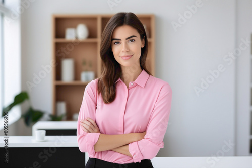 A minimalist office space with a professional ambiance, showcasing a young businesswoman wearing a pink shirt, radiating positivity and confidence, her arms crossed as she gazes into the camera.