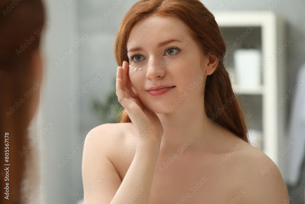 Beautiful woman with freckles and cream on her face near mirror in bathroom Beautiful woman with freckles and cream on her face near mirror in bathroom