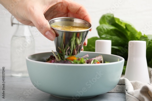 Woman pouring tasty vinegar based sauce (Vinaigrette) into bowl with salad at grey table, closeup photo