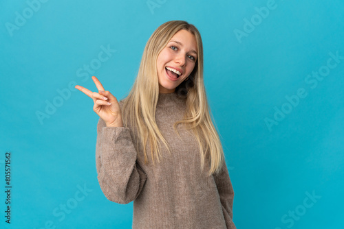 Young blonde woman isolated on blue background smiling and showing victory sign
