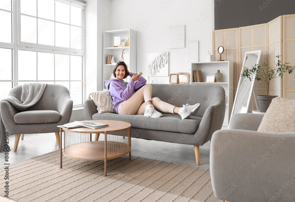Smiling young woman with air conditioner remote control lying on sofa in living room Smiling young woman with air conditioner remote control lying on sofa in living room