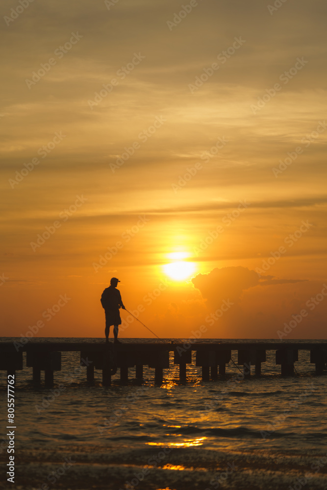 silhouette of a person fishing at the pier on the beach with sunset behind