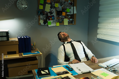 Exhausted investigator slumping in chair in cluttered office after long day of work