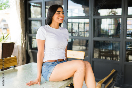 Cheerful young latina woman wearing a plain white t-shirt suitable for mockups, sitting outdoors at a cafe table, looking away