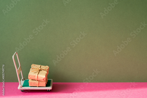 Trolley cart with gift boxes on pink and khaki green background. Parcel Delivery Concept