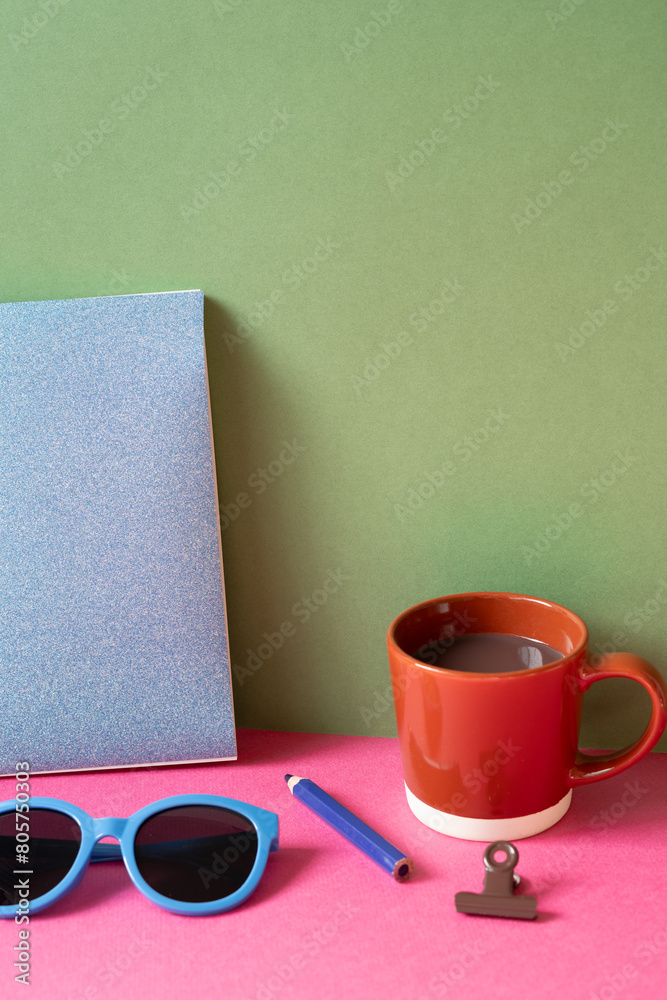 Desktop workspace. notebook, eyeglasses, colored pencil, clip, cup of hot chocolate on pink desk. khaki green wall background