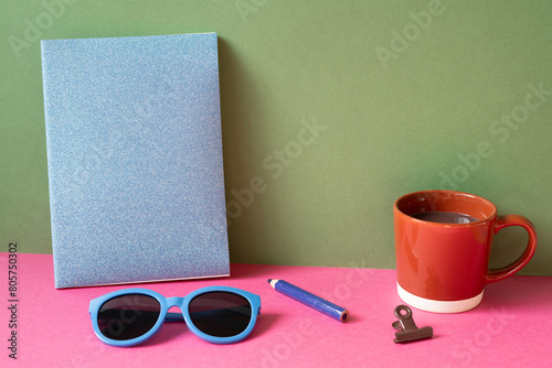 Desktop workspace. notebook, eyeglasses, colored pencil, clip, cup of hot chocolate on pink desk. khaki green wall background
