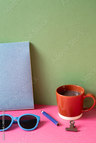 Desktop workspace. notebook, eyeglasses, colored pencil, clip, cup of hot chocolate on pink desk. khaki green wall background