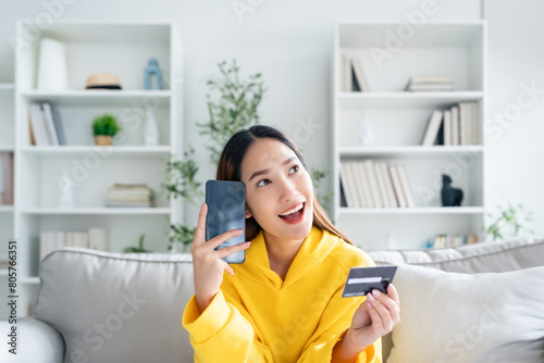 Young asian woman lying on sofa in living room  makes online banking payments through the internet from bank card on cell phone. Shopping online on mobile with credit card