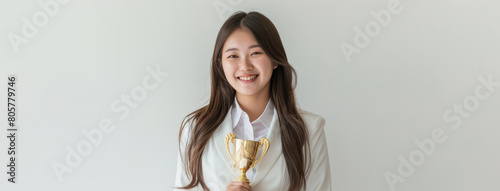 an Asian teenager clad in a pristine white suit and shirt proudly holds a gold trophy, symbolizing her dedication to academic success, against a white backdrop. photo