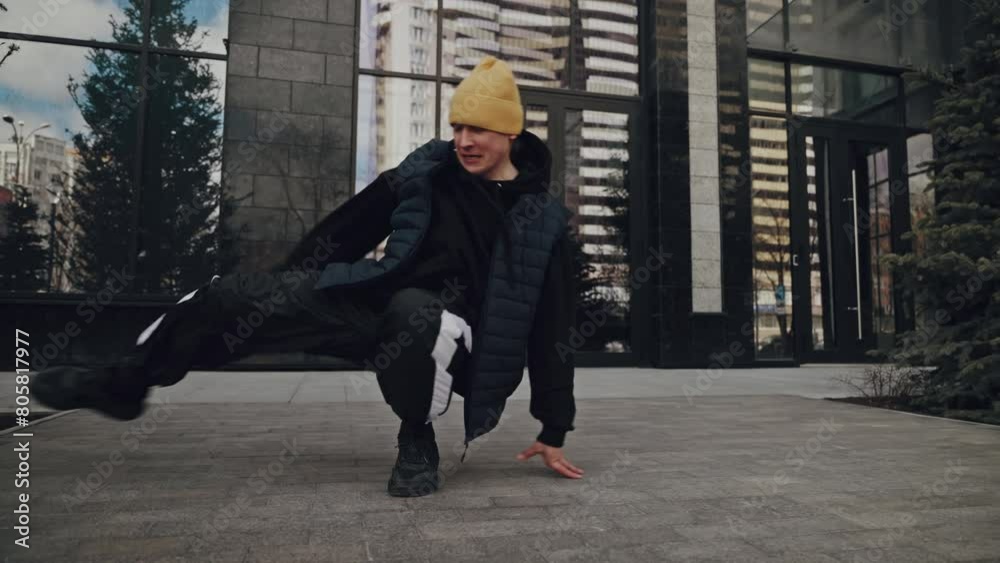 Person in yellow beanie cap sitting on ground with arm extended in front of a modern glass building, city reflections in the windows Person in yellow beanie cap sitting on ground with arm extended in front of a modern glass building, city reflections in the windows