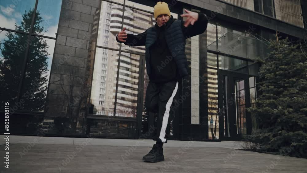 Person in yellow beanie cap and black jacket practicing urban dance moves in front of a modern glass building, with reflections of cityscape and sky in windows Person in yellow beanie cap and black jacket practicing urban dance moves in front of a modern glass building, with reflections of cityscape and sky in windows