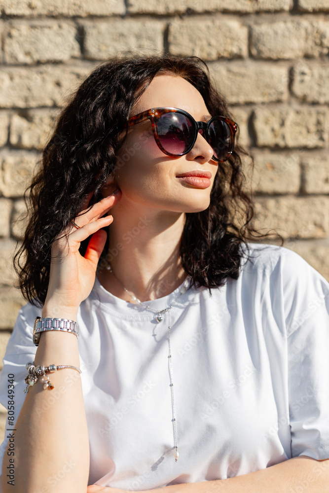 Summer female portrait of a beautiful fashion young woman model with curly hairstyle with sunglasses in a white t-shirt in the rays on the street Summer female portrait of a beautiful fashion young woman model with curly hairstyle with sunglasses in a white t-shirt in the rays on the street