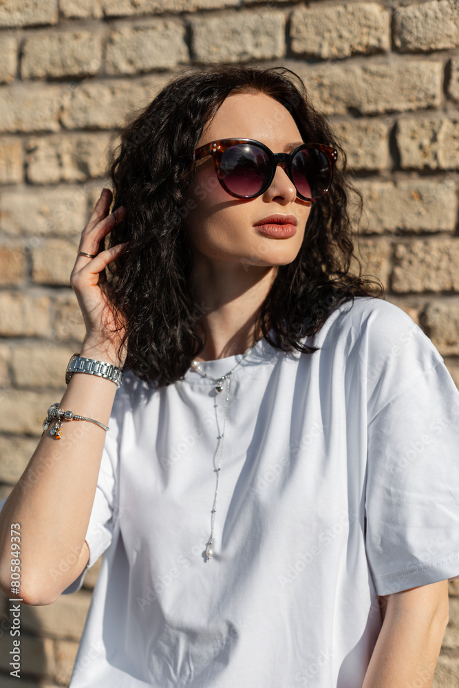 Stylish beautiful summer girl with curly hairstyle with sunglasses in a fashion white T-shirt near a brick wall on the street Stylish beautiful summer girl with curly hairstyle with sunglasses in a fashion white T-shirt near a brick wall on the street