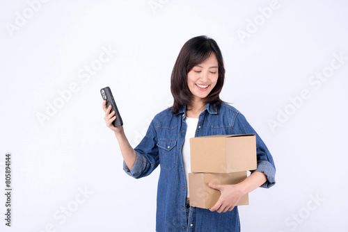 asian woman wearing denim jean holding parcel box while using mobile phone isolated on white studio background, Delivery courier and shipping service concept.