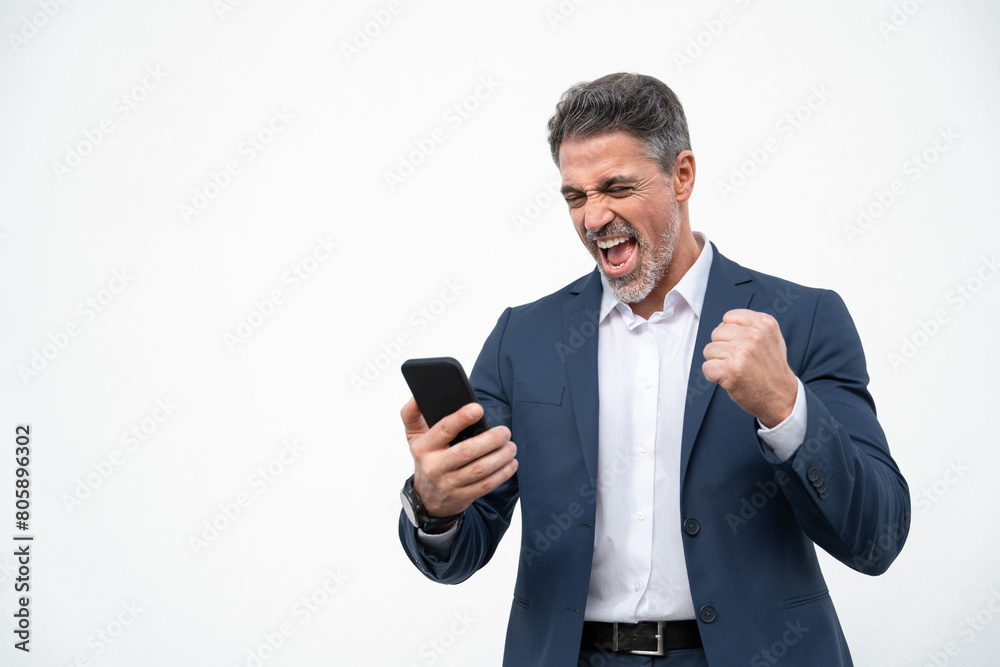 Happy delighted satisfied business man looking at mobile phone screen gesturing yes winning pose, isolated background. Overjoyed excited businessman in suit make winner gesture reading happy good news Happy delighted satisfied business man looking at mobile phone screen gesturing yes winning pose, isolated background. Overjoyed excited businessman in suit make winner gesture reading happy good news