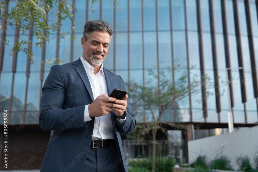 Smiling mature hispanic senior business man using smartphone cellphone at office building. Latin middle age male entrepreneur businessman holding mobile cell phone for trading outdoors. Copy space