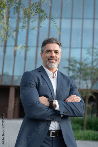 Vertical portrait older senior businessman professional ceo, manager, leader looking at camera at office building outdoors. Smiling confident latin hispanic mature business man standing crossed arms.