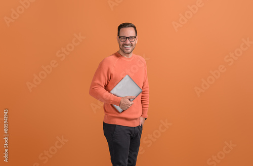 Portrait of young inspired businessman holding wireless computer and smiling on orange background