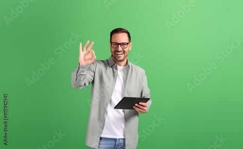 Portrait of young man showing OK sign and checking social media apps over tablet on green background
