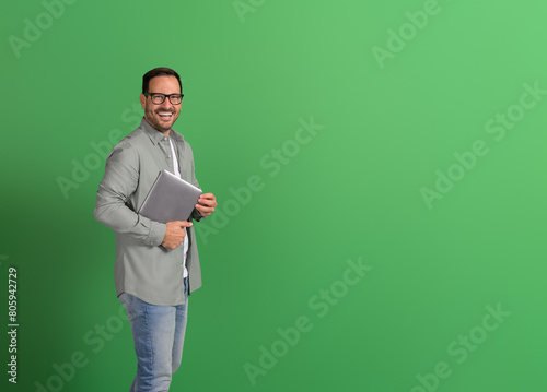 Portrait of young man in eyeglasses smiling and holding wireless laptop on isolated green background