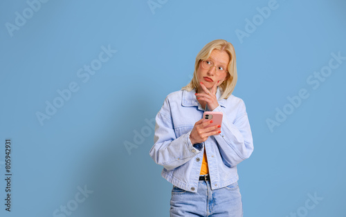 Young woman with hand on chin looking away thoughtfully while using mobile phone on blue background