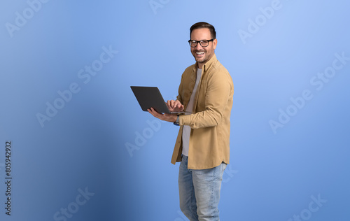 Portrait of cheerful young man in eyeglasses studying online over laptop on isolated blue background