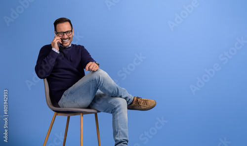 Happy male professional discussing over phone call while sitting on chair against blue background