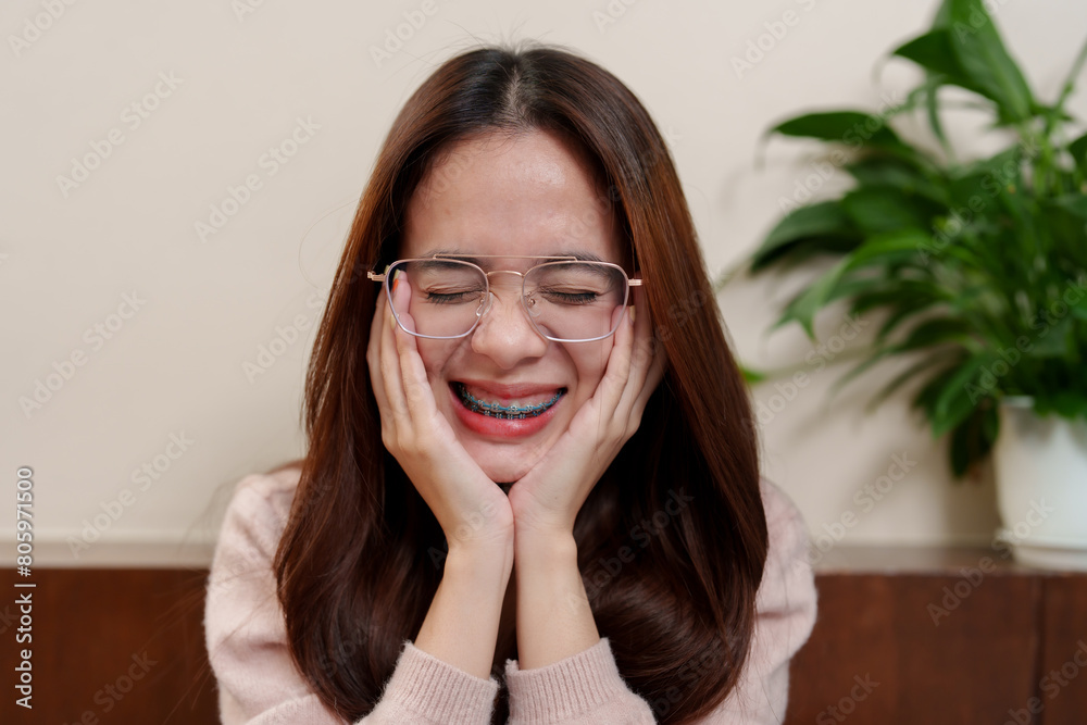 Adolescent with chestnut hair, light-framed spectacles, displaying exuberance, hands cupping cheeks, mouth open in delighted expression, orthodontic braces visible. Holiday happiness Asian teenagers Adolescent with chestnut hair, light-framed spectacles, displaying exuberance, hands cupping cheeks, mouth open in delighted expression, orthodontic braces visible. Holiday happiness Asian teenagers