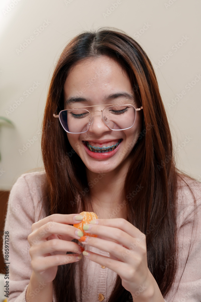 Youth in pastel sweater peeling orange with delight, glasses, braces, exudes comfort, simplicity of enjoying small pleasures on holiday. Holiday happiness Asian teenagers