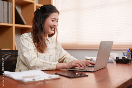 Smiling, Asian woman types on laptop, well-organized work desk, tablet, papers in home office. Joyful professional using computer, stationery neatly placed, comfortable workspace with window blinds.