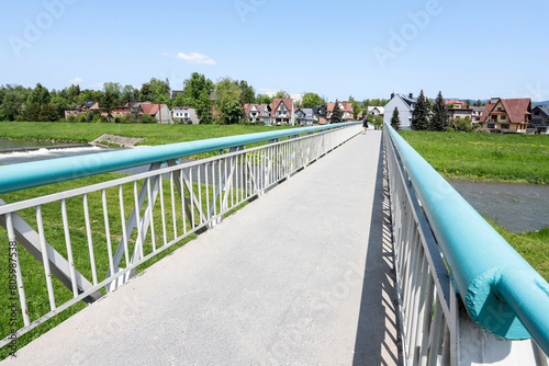 NOWY TARG, POLAND - MAY 27, 2023: Pedestrian bridge by the Dunajec River in Nowy Targ. Charming estates of single-family houses right by the water. photo