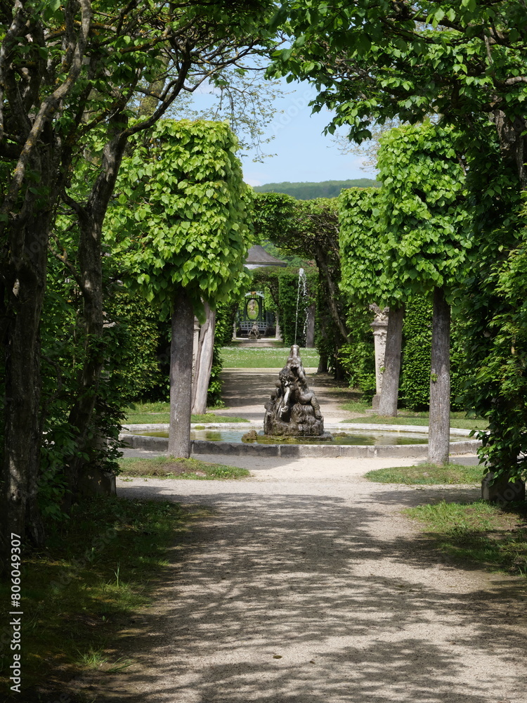 Romantischer Fontänenbrunnen mit einer Statue vor einem Laubengang zu einem Gartenpavillon in einer barocken Parkanlage