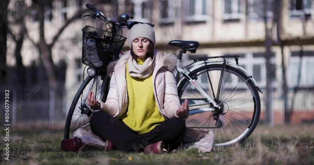 A peaceful scene of a young woman meditating outdoors next to her parked bicycle, enjoying a serene yoga session in an urban park setting.