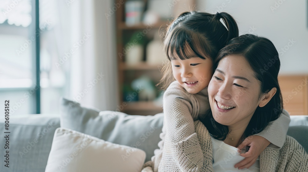 Asian woman sitting on the sofa with her daughter into hugging mom's shoulder, both smiling happy on living room. Asian woman sitting on the sofa with her daughter into hugging mom's shoulder, both smiling happy on living room.